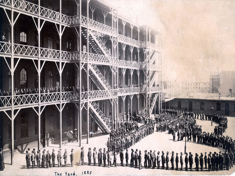 Hundreds of young boys, dressed in jackets and bowler hats, make a zig-zag line in the courtyard and up the stairs and on the mezzanine of one of the Catholic Protectory buildings, while a few Christian Brothers look on.